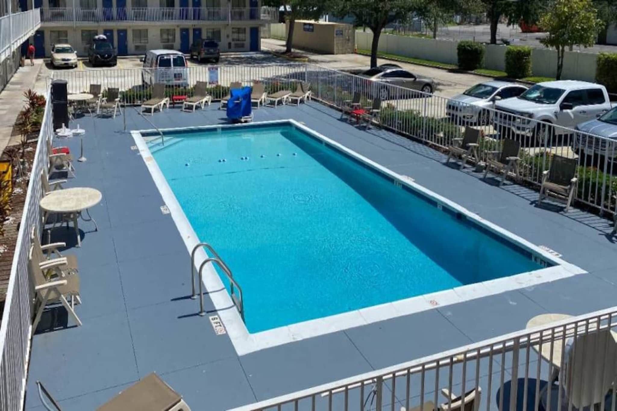 Ein rechteckiger Swimmingpool mit weißem Rand, umgeben von einem Metallzaun. Liegestühle und Tische befinden sich rund um den Pool.