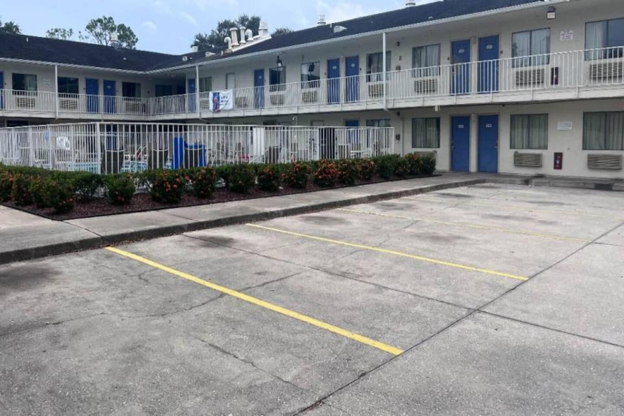 A motel with a parking lot marked with yellow lines, a fenced area with plants, and a row of blue doors on the building.