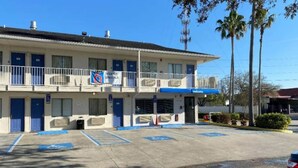 Um motel de dois andares com esquema de cores azul e branco, estacionamento com vagas para deficientes e um letreiro com 'Promoções Semanais'.