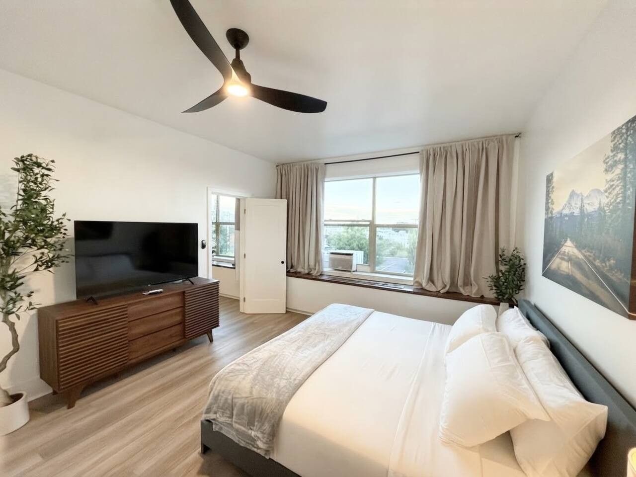 Un dormitorio con cama, televisor, ventilador de techo, ventana con cortinas y una planta.