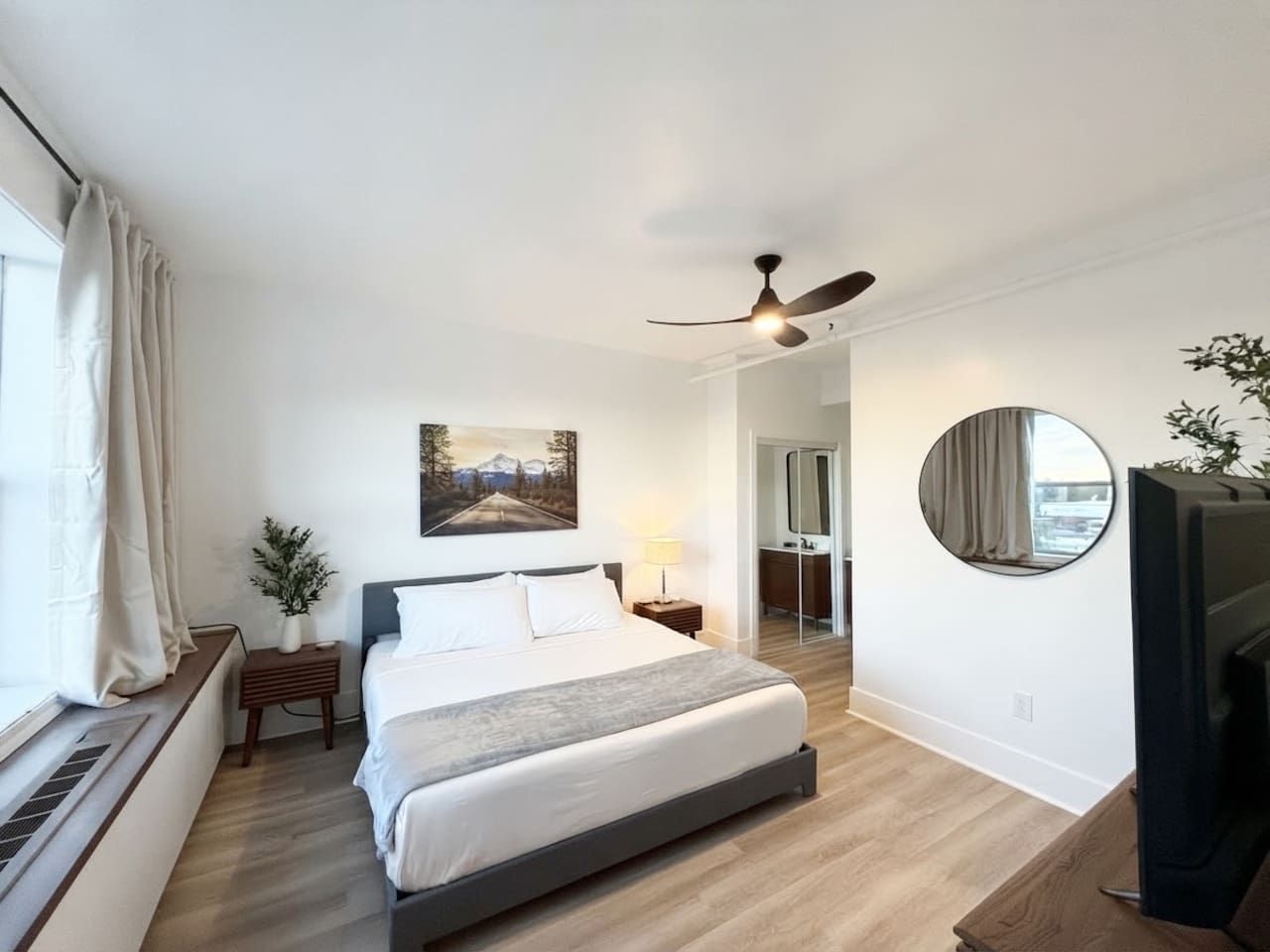 A bedroom with a bed, bedside table, lamp, ceiling fan, and a round mirror.