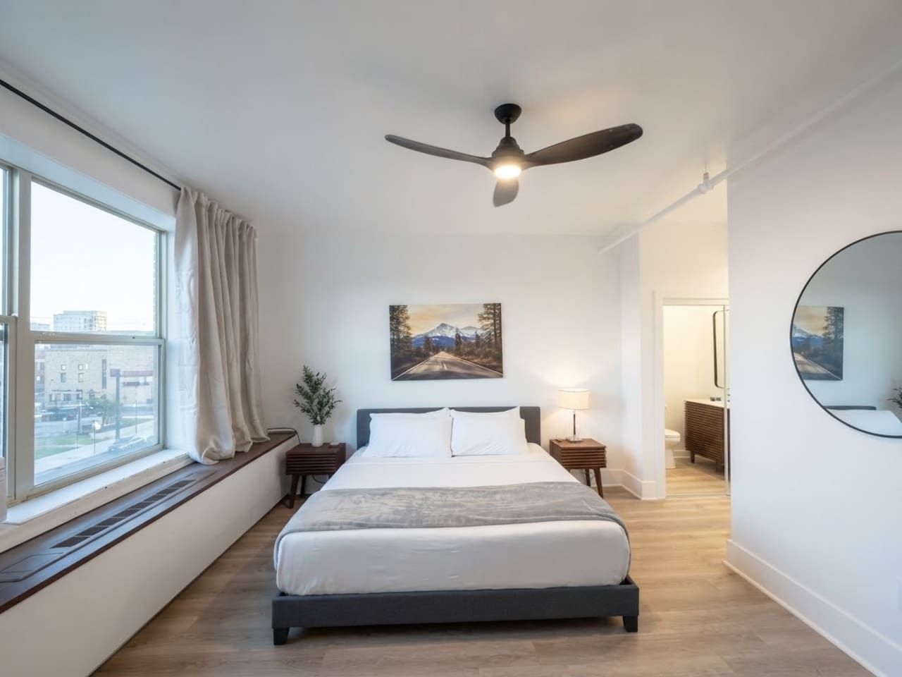 Een slaapkamer met een bed, een plafondventilator, een raam met gordijnen, een schilderij aan de muur en een ronde spiegel.
