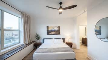 Een slaapkamer met een bed, een plafondventilator, een raam met gordijnen, een schilderij aan de muur en een ronde spiegel.