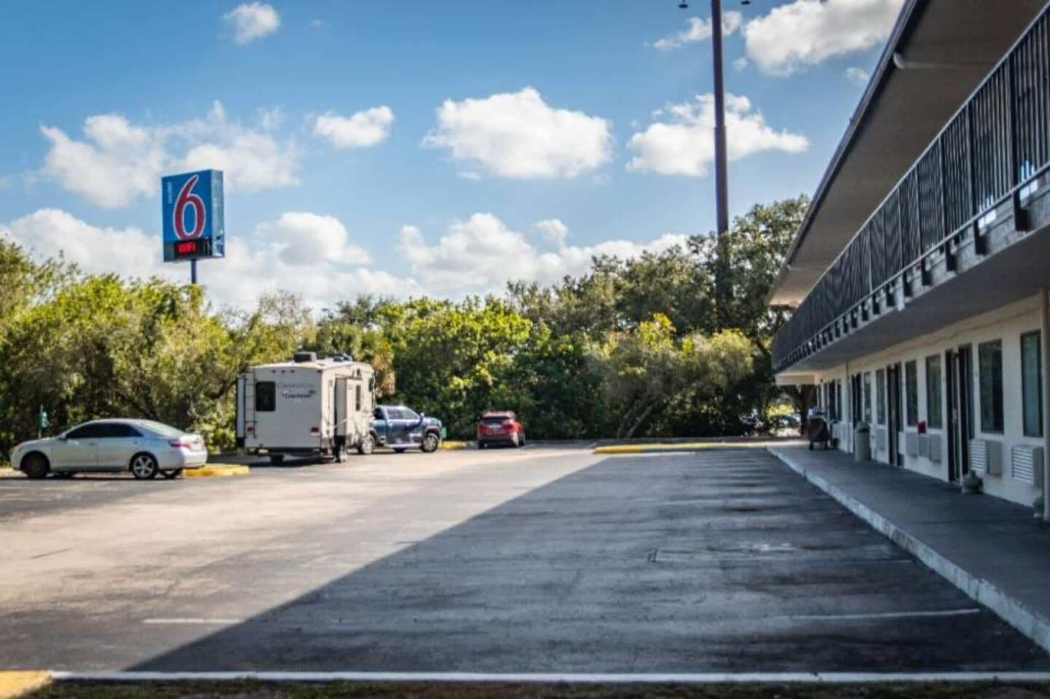 Ein Parkplatz mit Wohnmobilen und Autos, ein Gebäude mit einer Beschilderung mit der Zahl '6' und Bäume im Hintergrund.