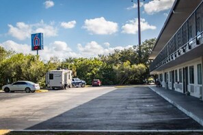 Un estacionamiento con vehículos recreativos y automóviles, un edificio con un letrero '6' y árboles al fondo.