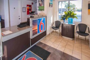 A reception area with a desk, chairs, a large window, and a sign indicating the number 6. - Motel 6 Ft. Pierce, Fl_1 Queen Bed | Ada (Fort Pierce)