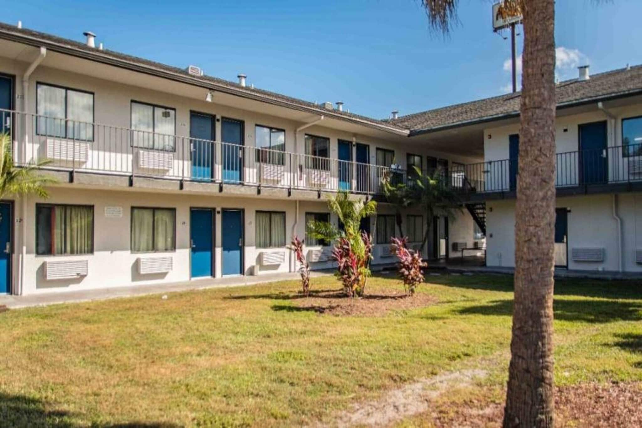 A multi-story motel with a flat roof, multiple balconies, and a small lawn area with a palm tree.