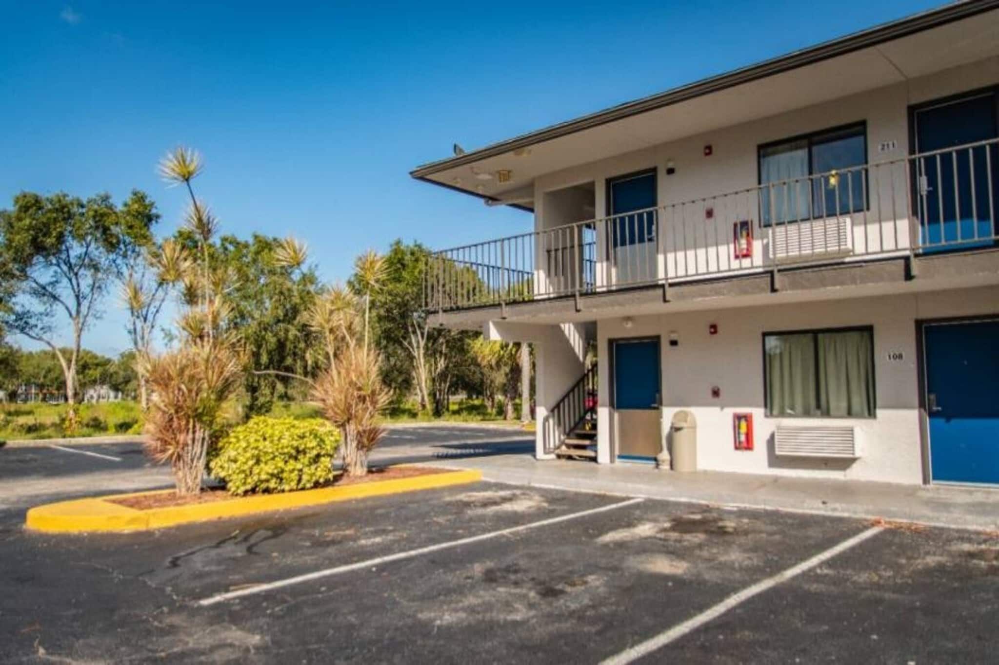 A two-story motel with a balcony on the second floor, a parking lot with marked spaces, and a small garden area with palm trees.