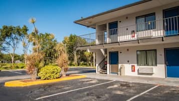 A two-story motel with a balcony on the second floor, a parking lot with marked spaces, and a small garden area with palm trees.