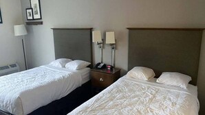 Duas camas de solteiro com cabeceiras, uma mesa de cabeceira com telefone e relógio, e uma luminária fixada na parede.
