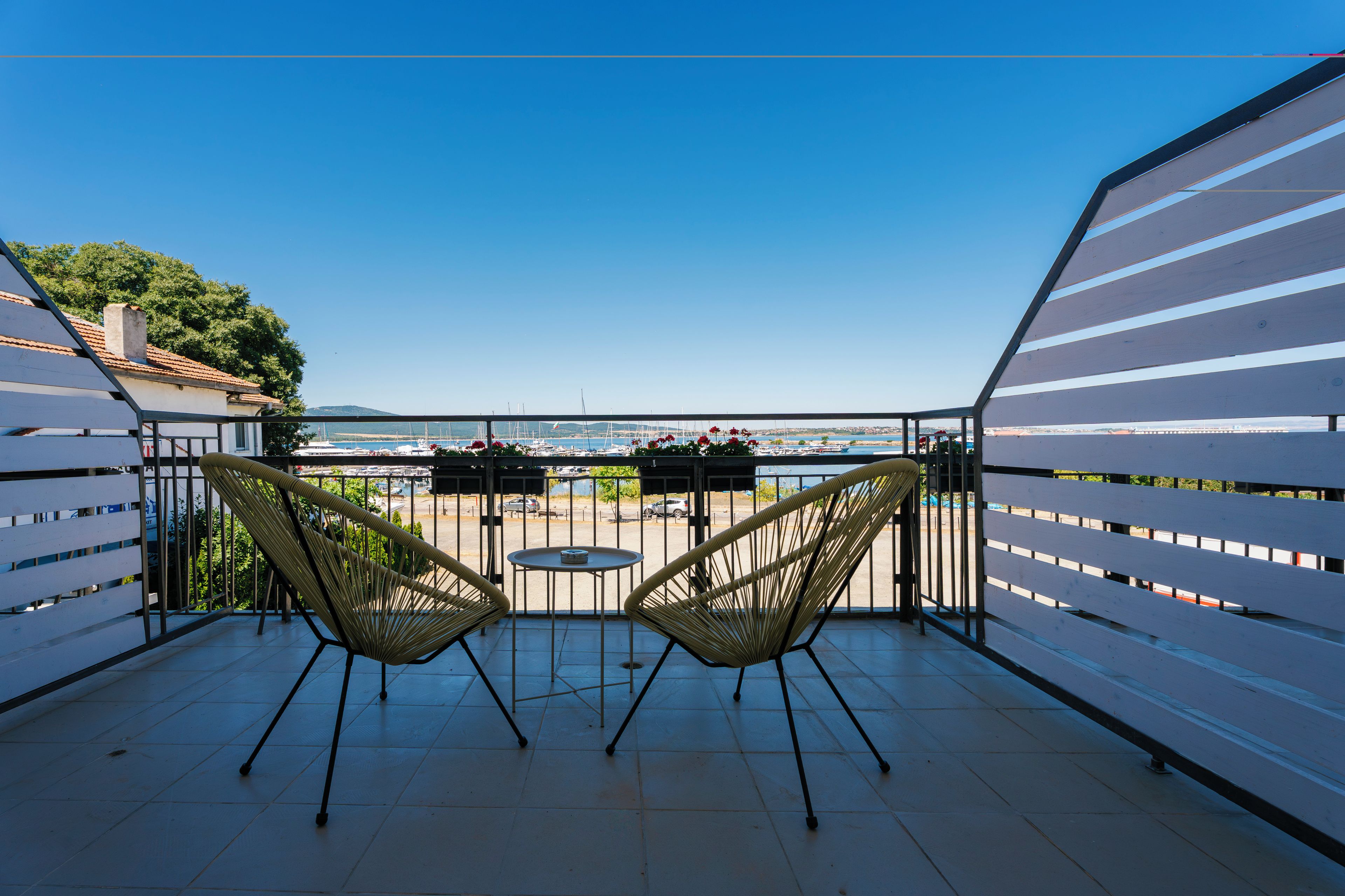 A balcony with two wicker chairs and a small round table, overlooking a marina with boats and a clear blue sky.