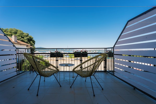 A balcony with two wicker chairs and a small round table, overlooking a marina with boats and a clear blue sky. - Hotel Sozopol 99 (Sozopol)