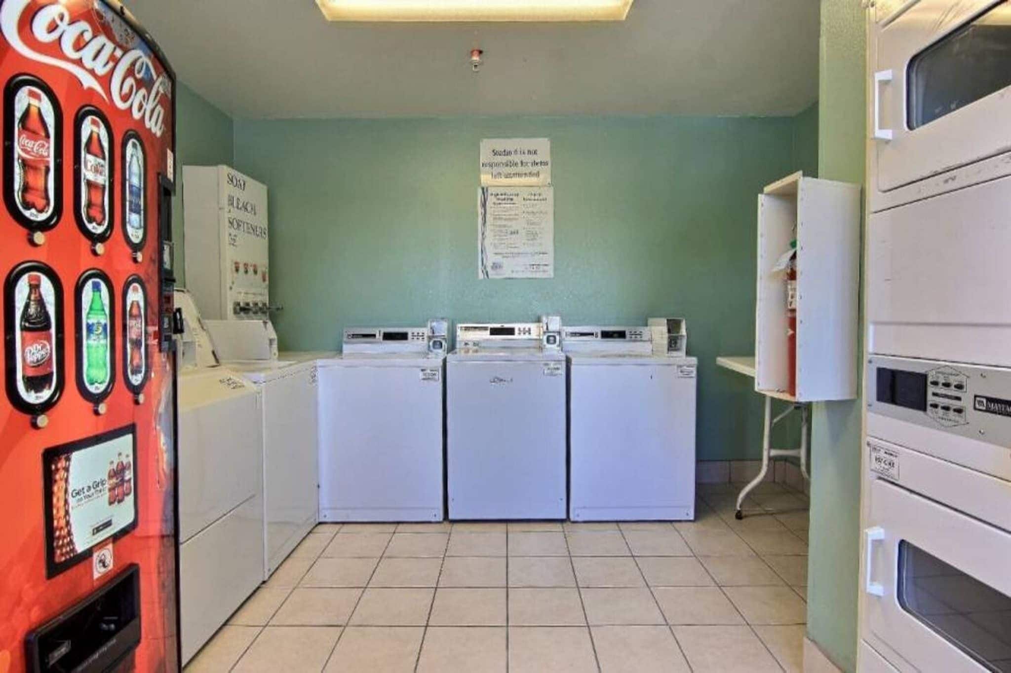 A laundromat with three washers and dryers, a Coca-Cola vending machine, and a sign on the wall.