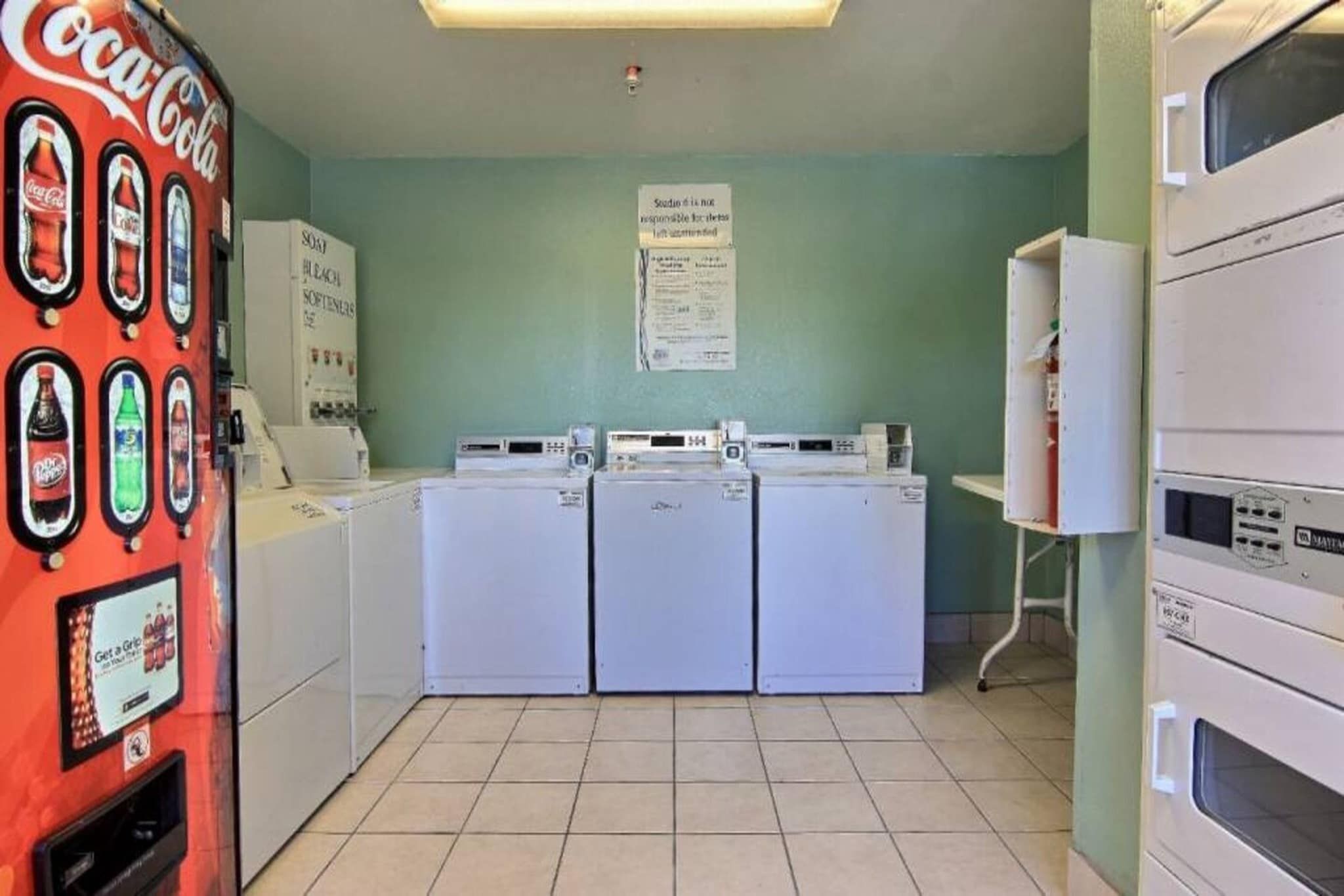 Una lavanderia self-service con tre lavatrici e asciugatrici, una macchina distributrice di Coca-Cola e un cartello appeso al muro.