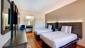 A hotel room with two beds, a wooden floor, a desk with a mirror, and a small bathroom. - 2 Full Beds | Non-Smoking (Gainesville)