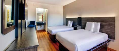 A hotel room with two beds, a wooden floor, a desk with a mirror, and a small bathroom.