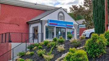 A building with a 'WELCOME' sign, a covered entrance, and a landscaped area with shrubs and a small hill.