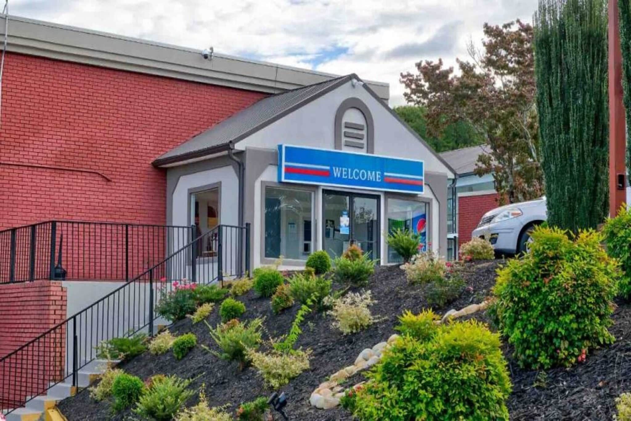 A building with a 'WELCOME' sign, a covered entrance, and a landscaped area with shrubs and a small hill.