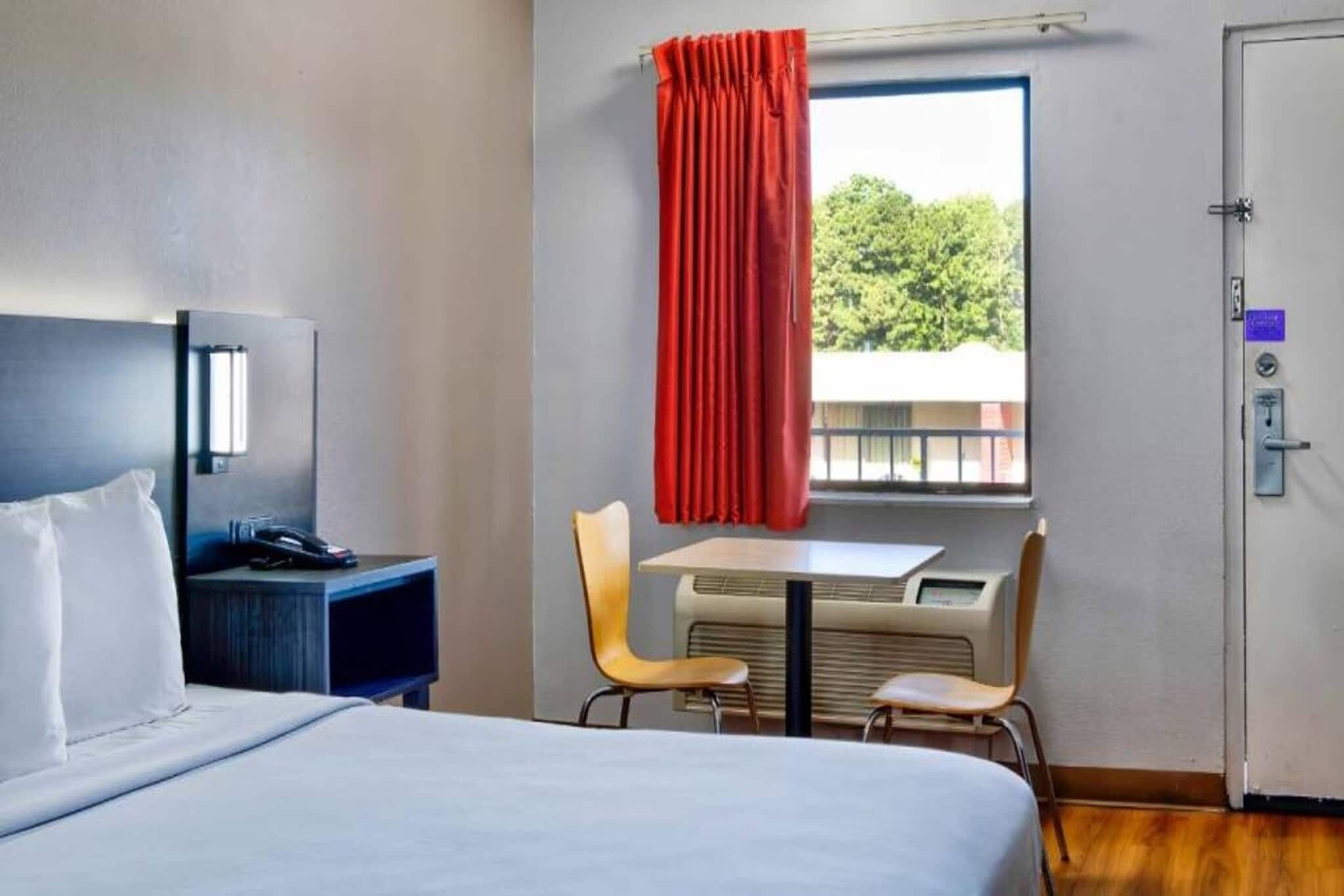A hotel room with a bed, a desk, a chair, a table, an air conditioning unit, a window with curtains, and a door.