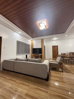 A modern living room with a sectional sofa, a TV, wooden flooring, and a ceiling light fixture. - Casita Nela: Cozy 3BR Private Haven near Tagaytay (Silang)