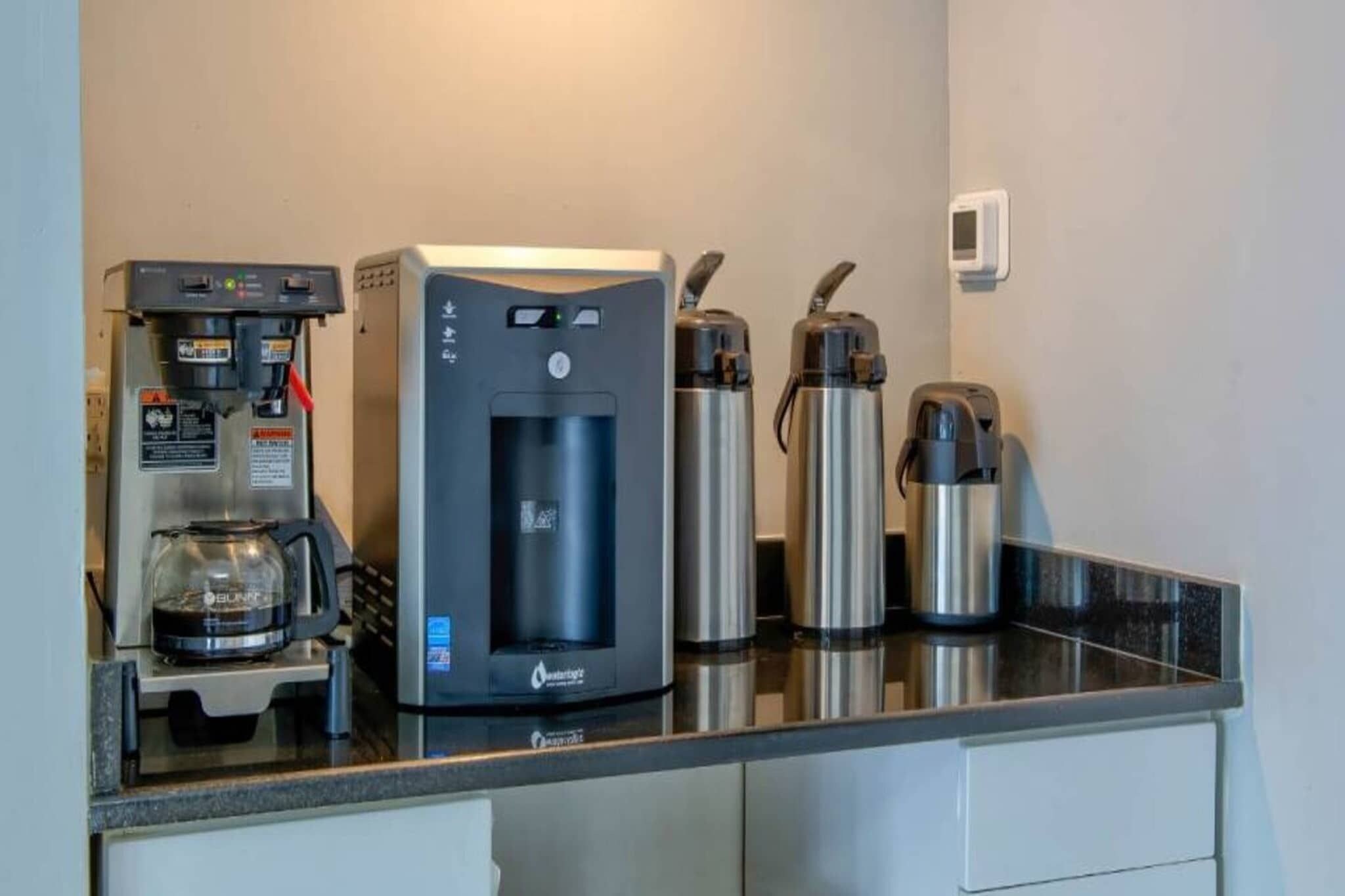 A kitchenette with a water dispenser, a coffee machine, and two stainless steel thermos cups.