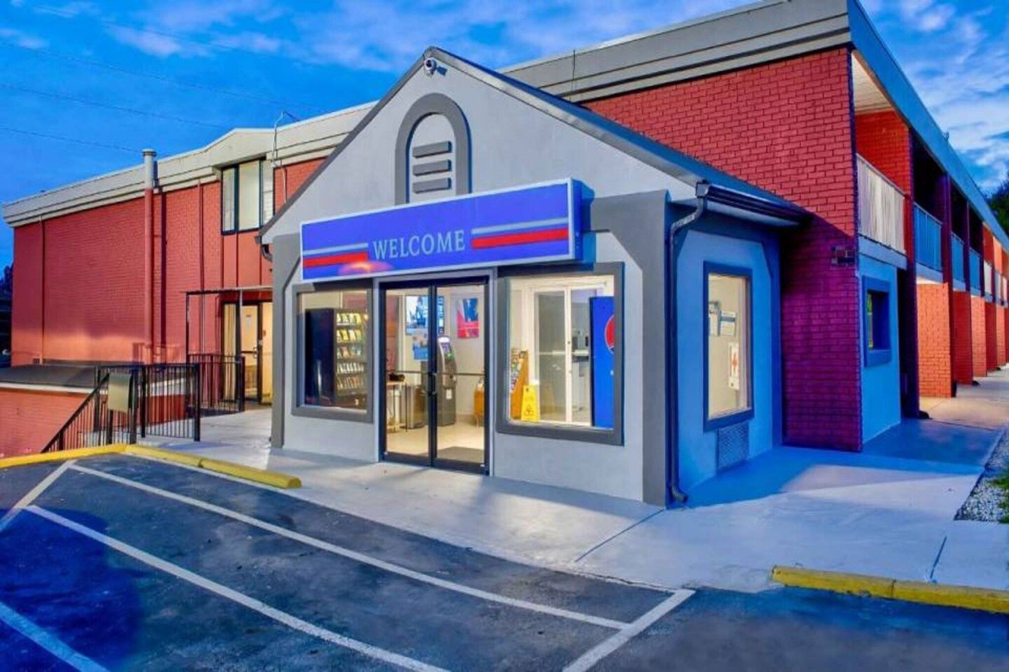 A building with a 'WELCOME' sign, a parking lot, and a red brick wall.