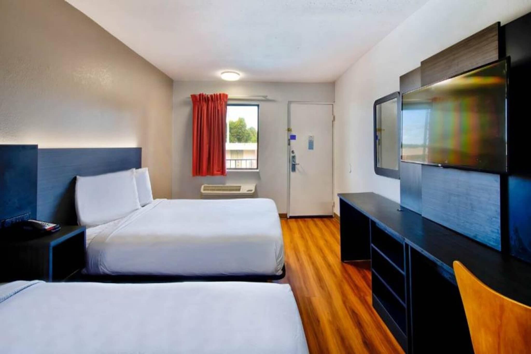 A hotel room with two beds, a TV, a desk, and a window with curtains.