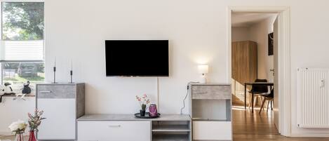 Una sala de estar moderna con un televisor de pantalla plana montado en la pared, un comedor con una mesa y sillas, y una cocina con armarios y una encimera.