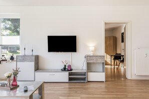 Un salon moderne doté d’un téléviseur à écran plat fixé au mur, d’un coin repas avec une table et des chaises, et d’une cuisine équipée de placards et d’un plan de travail.