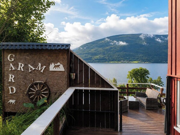 A wooden deck with a view of a lake and mountains, a rustic building with a sign reading 'GARMD', and outdoor furniture.