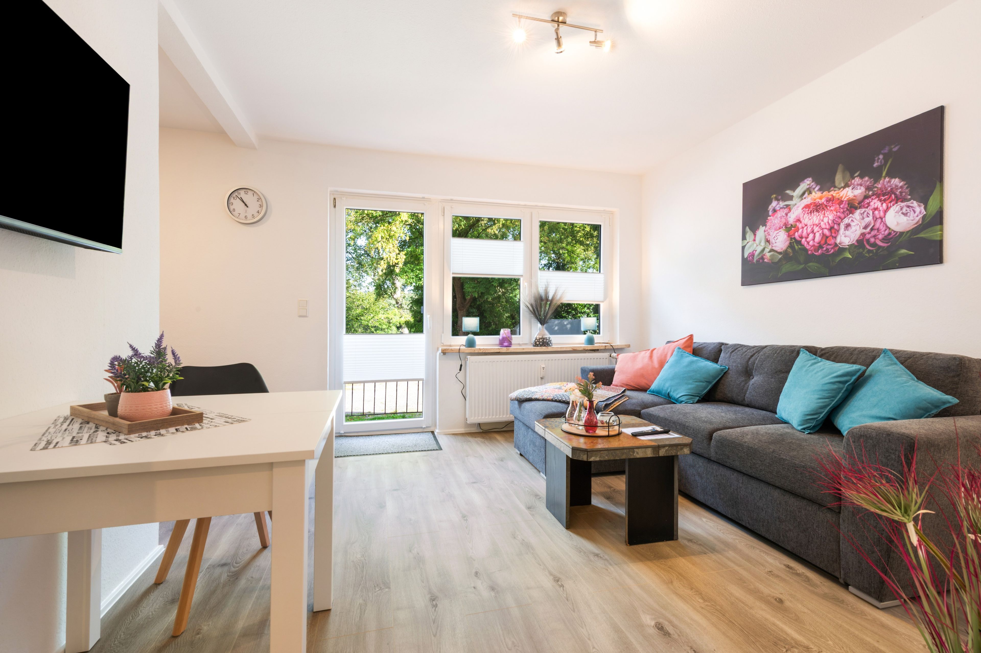 Ein modernes Wohnzimmer mit grauem Sofa, einem Esstisch und einem durch ein großes Fenster sichtbaren Küchenbereich.