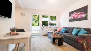 Ein modernes Wohnzimmer mit grauem Sofa, einem Esstisch und einem durch ein großes Fenster sichtbaren Küchenbereich.