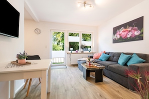 Ein modernes Wohnzimmer mit grauem Sofa, einem Esstisch und einem durch ein großes Fenster sichtbaren Küchenbereich.