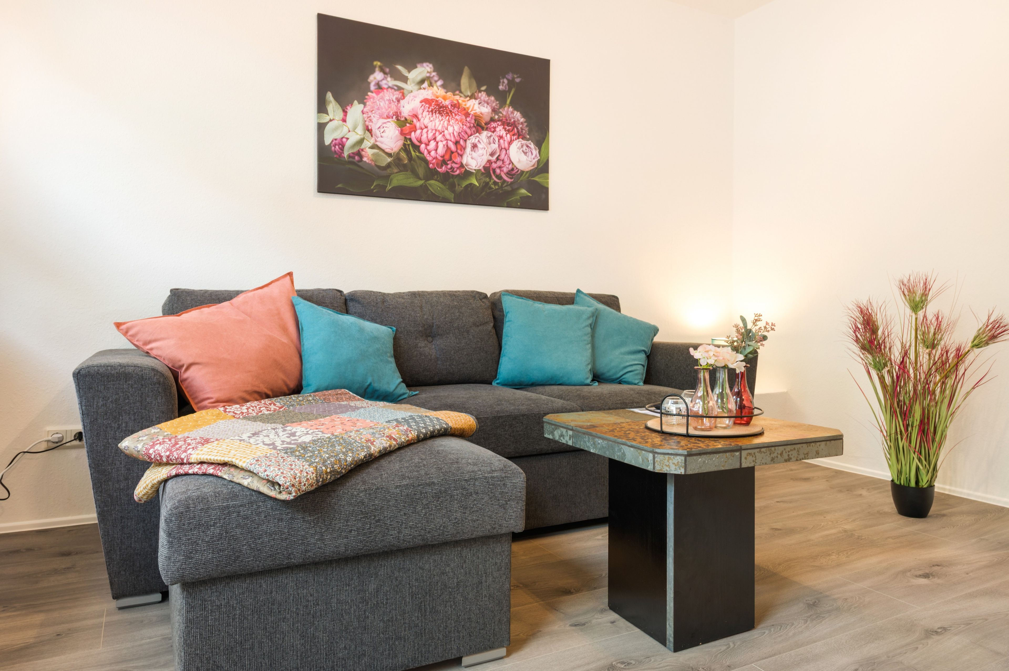 Ein Wohnzimmer mit einem grauen Sofa, bunten Kissen, einer gemusterten Decke und einem kleinen Beistelltisch mit einem Blumenstrauß.