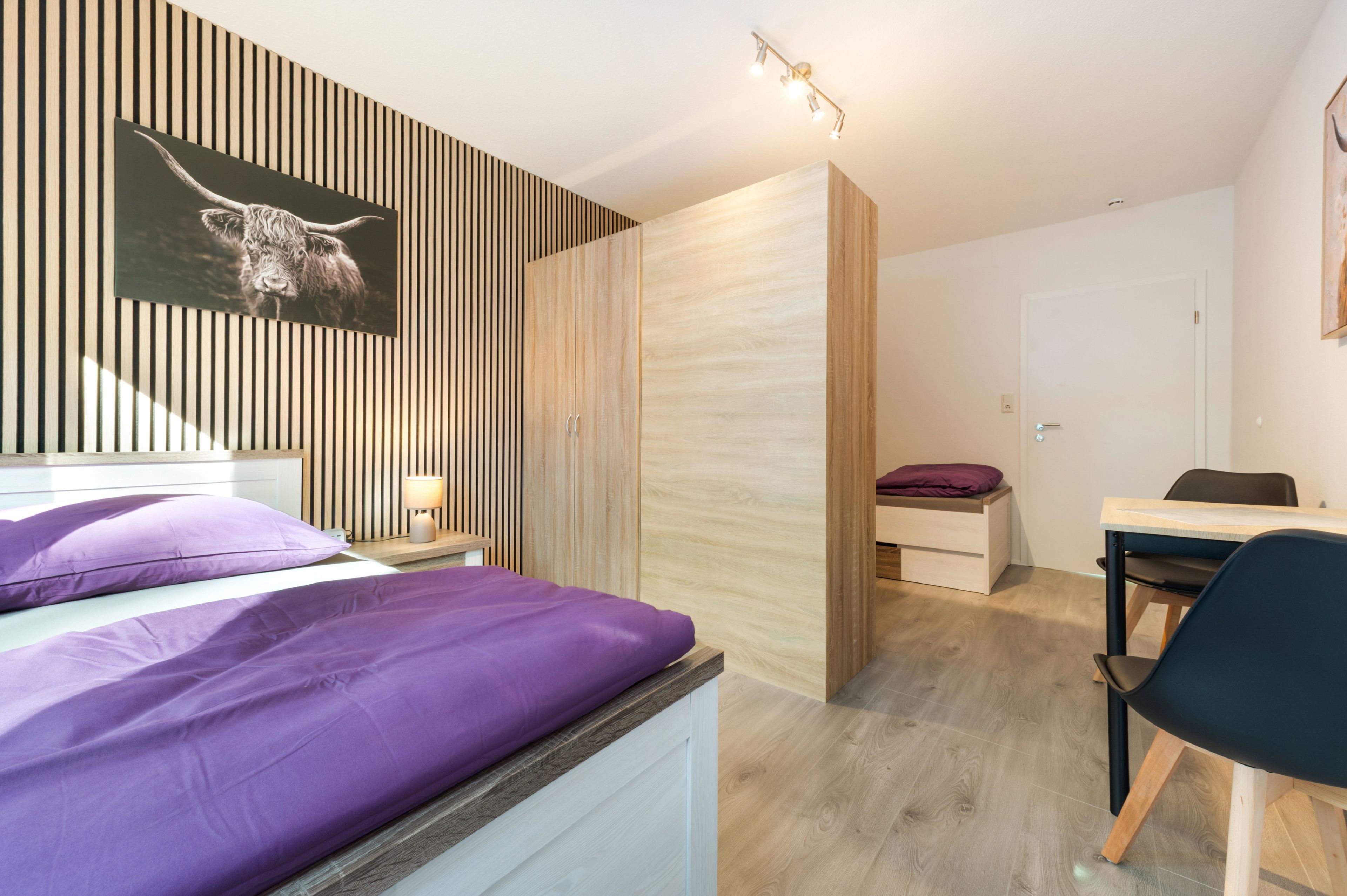 Een moderne slaapkamer met een bed, een bureau en een stoel. Er is een houten garderobe en een wandlamp.