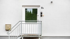 Edifício branco com porta marcada '8b', uma pequena escadaria externa e um sistema de interfone de segurança.
