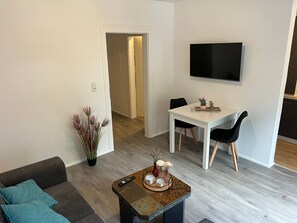 A modern living room with a sofa, coffee table, dining table, chairs, and a flat-screen TV mounted on the wall. - Apartment "Zum Waldblick 3" with Shared Garden and Wi-Fi (Wulsbüttel )