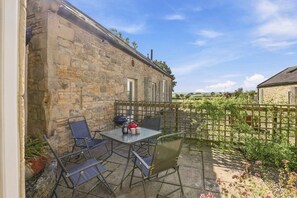 A stone building with a patio area featuring a table and chairs, a small garden with a fence, and a clear sky. - The Old Woodworker’s Cottage - North East Escapes (Great Whittington)