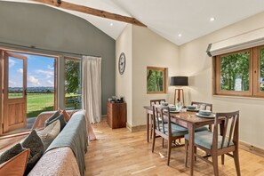 Ein Zimmer mit Esstisch für vier Personen, einer Couch mit Kissen, einer Lampe und Blick auf eine grüne Landschaft durch große Fenster.