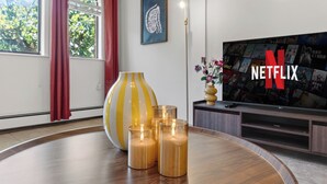 Ein modernes Wohnzimmer mit einem großen Flachbildfernseher, auf dem das Netflix-Logo zu sehen ist, einem runden Holztisch mit Kerzen, einer dekorativen Vase und einem Fenster mit Vorhängen.