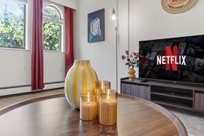 Uma sala de estar moderna com uma televisão de tela plana grande exibindo o logotipo da Netflix, uma mesa redonda de madeira com velas, um vaso decorativo e uma janela com cortinas.