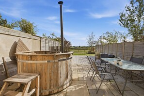 Een terras buiten met een houten jacuzzi, een tafel met stoelen en een barbecue.