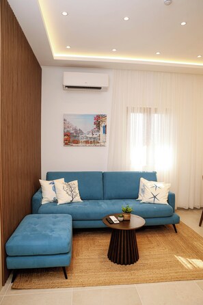 Ein modernes Wohnzimmer mit einem blauen Sofa, einem passenden Hocker, einem runden Couchtisch und einem Gemälde einer Küstenlandschaft.