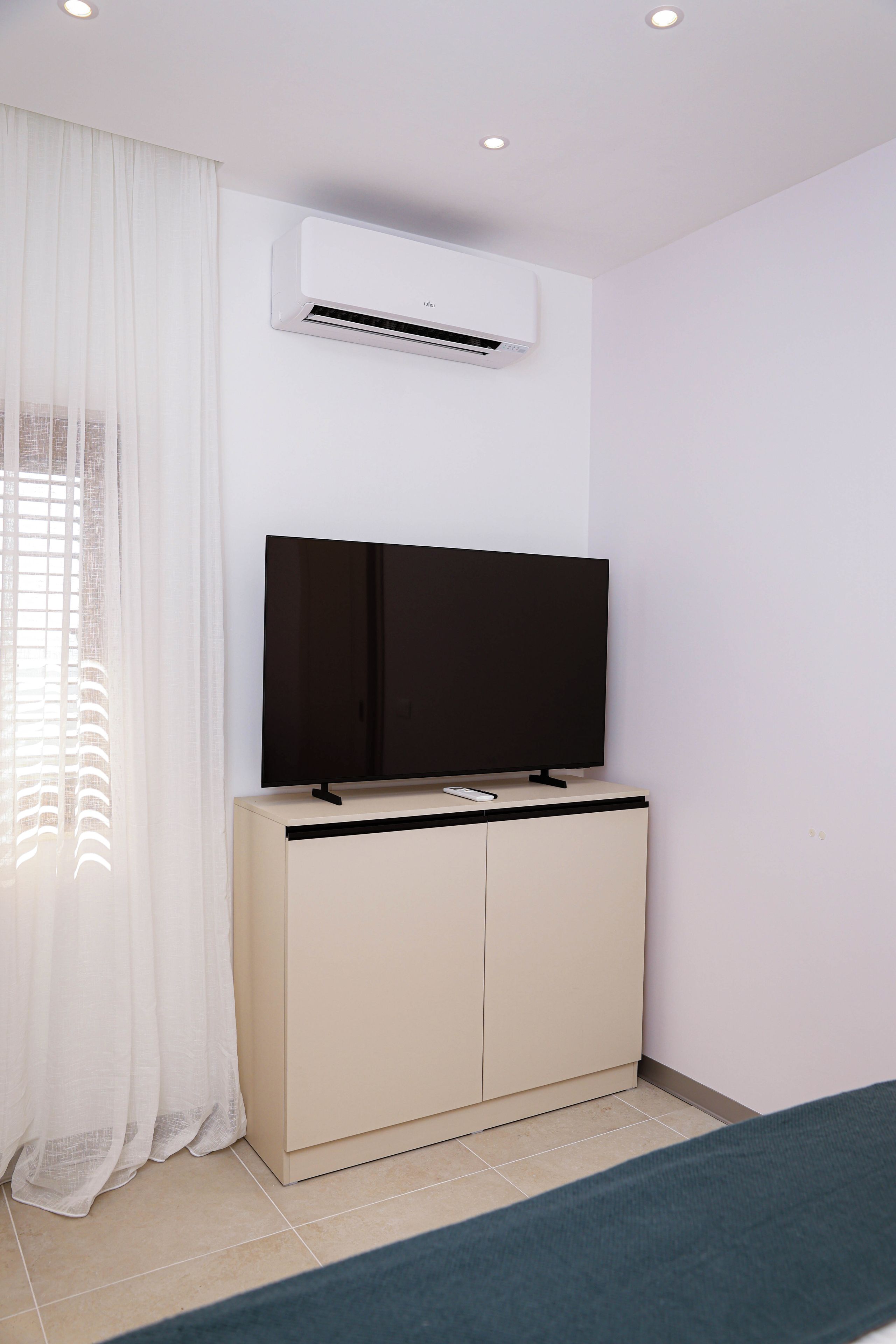 A room with a flat-screen TV on a cabinet, an air conditioning unit on the wall, and a window with sheer curtains.