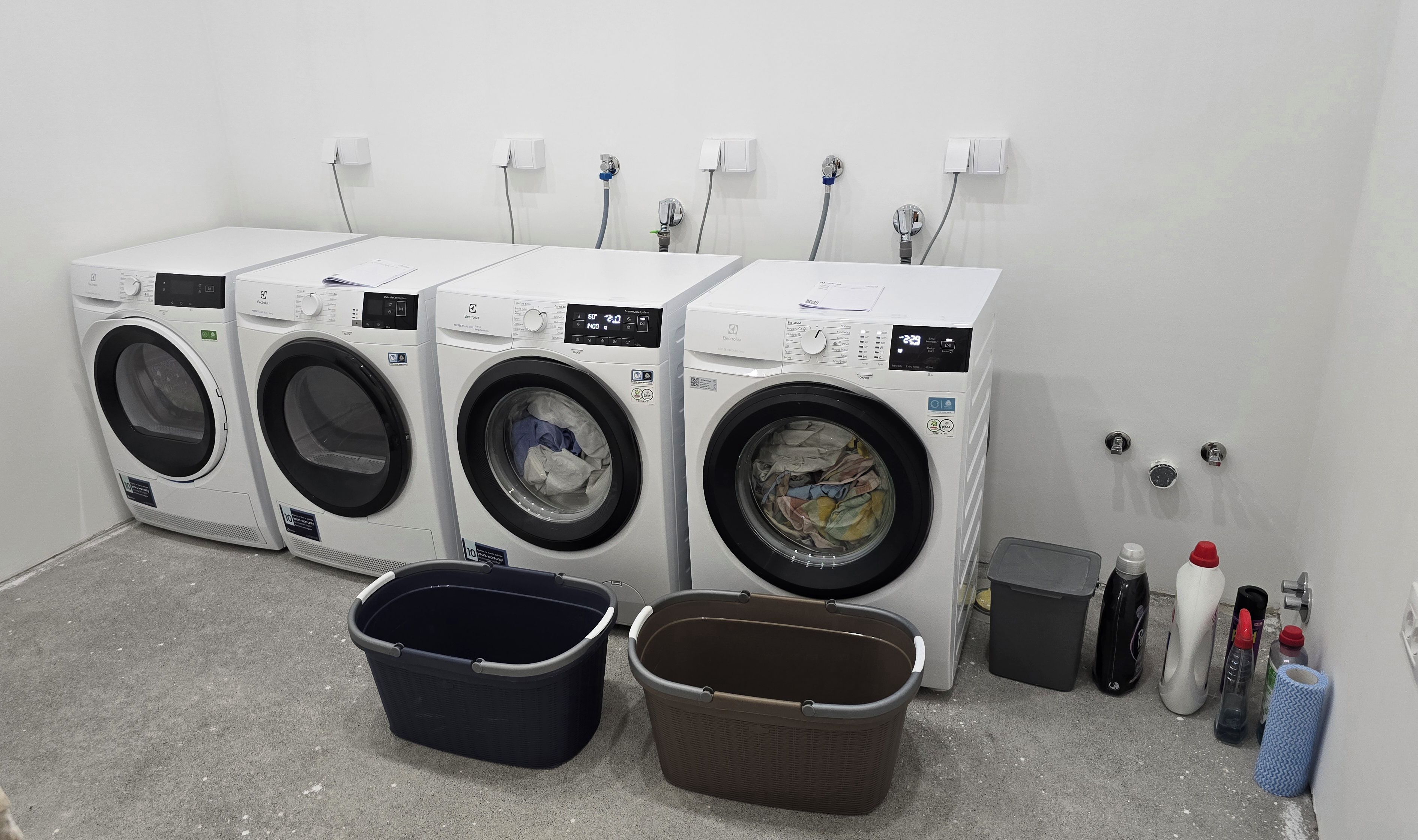 Une rangée de quatre machines à laver à chargement frontal dans une buanderie, avec des paniers et des produits de nettoyage.