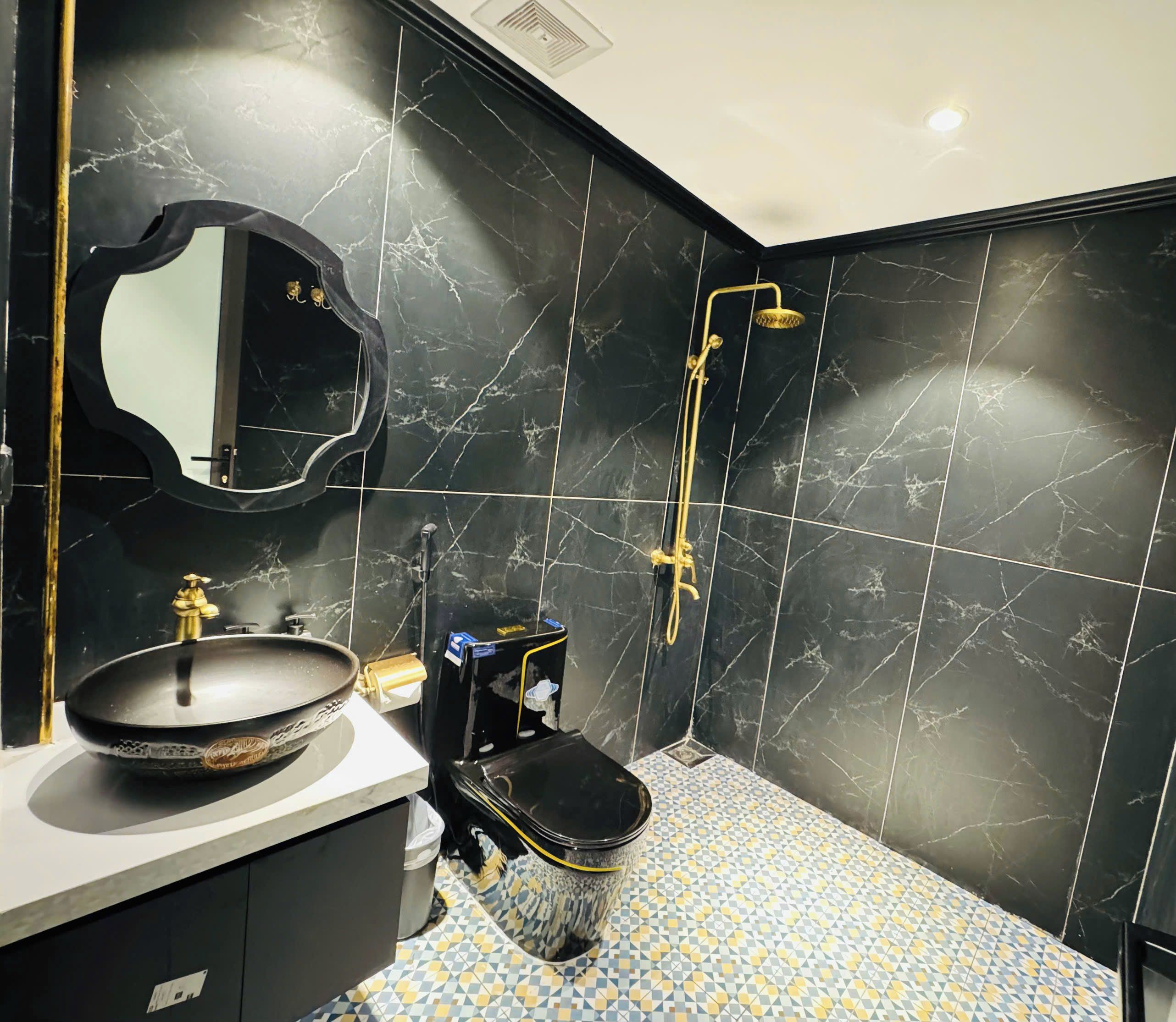 A bathroom with a black marble wall, a round sink, a mirror, a shower with a golden fixture, and a patterned floor.