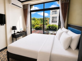 Habitación de hotel con una cama grande, una mesita de noche con lámpara, televisión y vistas a un balcón con plantas y un edificio.
