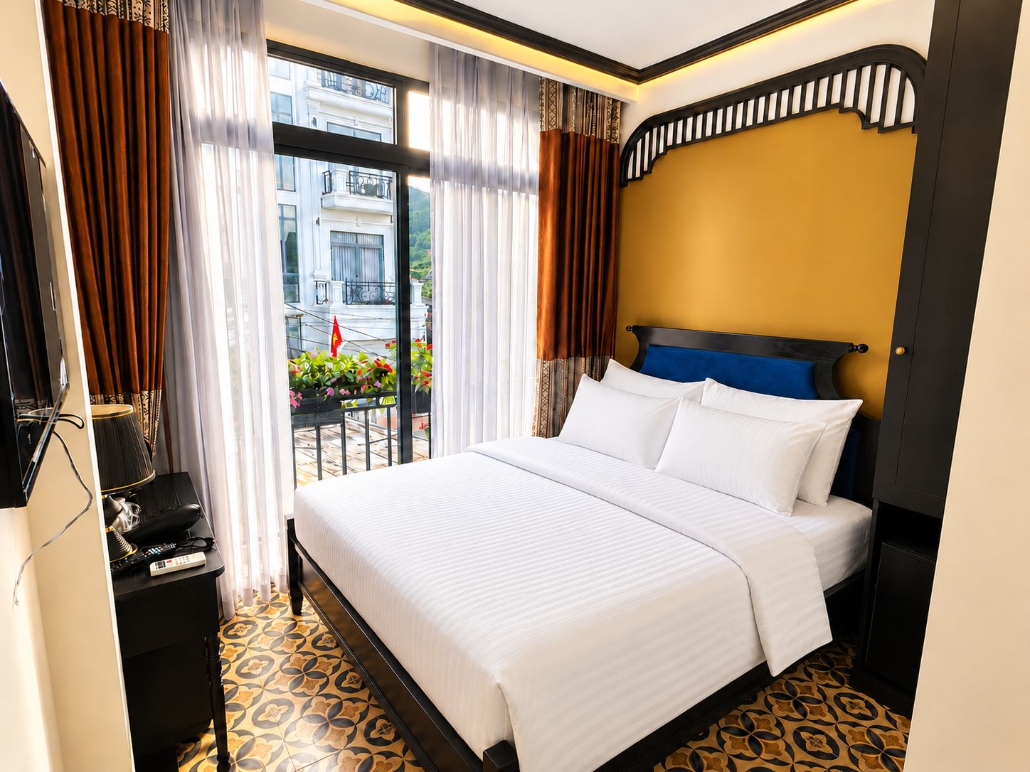 Habitación de hotel con cama, escritorio con teléfono y vista a un balcón con plantas y una bandera.