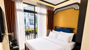 Habitación de hotel con cama, escritorio con teléfono y vista a un balcón con plantas y una bandera.