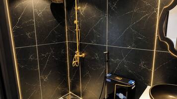 Un baño con pared de mármol negro, ducha con rociador dorado y lavamanos negro.